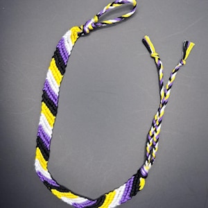 Könnte beinhalten: Ein Freundschaftsarmband in Chevron-Muster in den Farben Lila, Gelb und Schwarz.