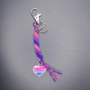 Può includere: Un portachiavi con un braccialetto dell'amicizia rosa, blu e viola con un ciondolo a forma di cuore con i colori della bandiera del pride bisessuale.