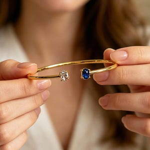 Può includere: Un bracciale aperto in oro con due pietre di forma ovale alle estremità. Una pietra è una gemma trasparente e incolore, l'altra è una gemma di colore blu intenso simile allo zaffiro. Il bracciale ha una finitura metallica liscia e lucida.