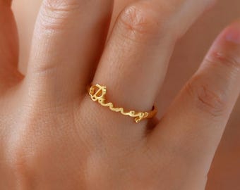 Op de afbeelding: Een goudkleurige ring met de naam "Darney" in cursief schrift. De ring heeft een klein kroontje aan het begin van de naam. De ring wordt aan een vinger gedragen.