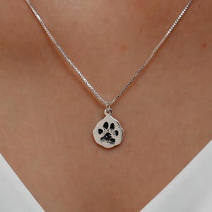 Puede incluir: Un collar de plata con un pequeño colgante en forma de pata. El colgante presenta un diseño de huella de pata negra. El collar se lleva sobre una parte superior blanca.