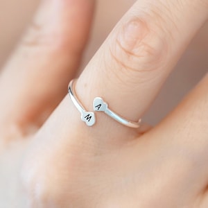Könnte beinhalten: Ein silberfarbener offener Ring mit zwei kleinen herzförmigen Enden. Ein Herz ist mit dem Buchstaben A und das andere Herz mit dem Buchstaben M graviert. Das Band ist dünn und metallisch und so gestaltet, dass es den Finger teilweise umschließt.