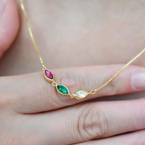 Puede incluir: Un delicado collar de oro con tres piedras preciosas de talla marquesa en rojo, verde y blanco. Las piedras están engastadas en un bisel de oro y están conectadas por una fina cadena de oro. El collar está siendo sostenido por una persona.