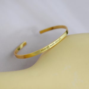 Puede incluir: Una pulsera de brazalete dorada con el texto grabado "Adversity builds strength". La pulsera es curva y abierta, diseñada para deslizarse sobre la muñeca. La joya se exhibe sobre una superficie amarillo claro.