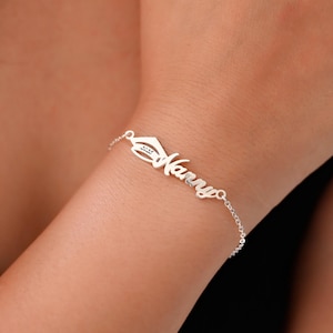 May include: A silver bracelet inscribed with the name "Nanny" in cursive. The bracelet includes a charm with a hat design. The bracelet is displayed on a wrist against a dark backdrop.