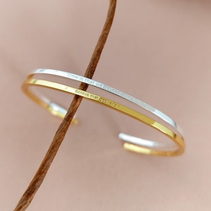 May include: Two open cuff bracelets, one gold and one silver, are shown. The gold bracelet has the text "Believe that god is fair" and the silver bracelet has the text "Time is that we'll not come late".