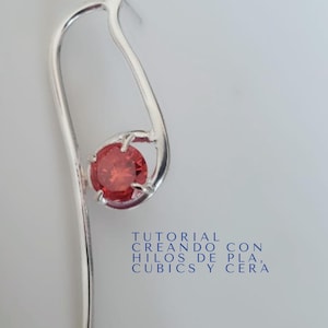 Puede incluir: Un pendiente plateado con un diseño curvo, con una gema roja redonda. El pendiente tiene un poste para la inserción y un lazo decorativo. La imagen también incluye el texto "JOYERÍA EXPERIMENTAL" y "TUTORIAL CREANDO CON HILOS DE PLA, CUBICS Y CERA".