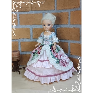 Könnte beinhalten: Eine Porzellanpuppe, die ein hellblaues und rosa Kleid mit Blumendruck und Spitzenbesatz trägt. Das Kleid hat einen gerüschten Rock und ein tailliertes Oberteil. Die Puppe hat blonde Haare und blaue Augen.