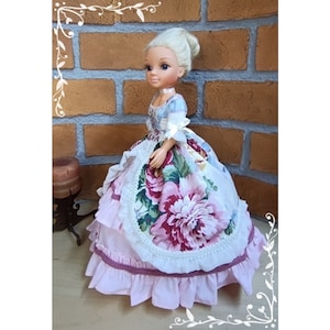 May include: A porcelain doll wearing a white and pink dress with floral print. The dress has a lace trim and multiple layers of ruffles. The doll has blonde hair and blue eyes.