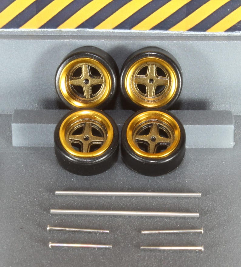 May include: Four black model car wheels with gold rims and a cross-shaped spoke design. Several silver metal rods and pins are also included. The wheels are arranged in a square formation, suggesting a set for a miniature vehicle.