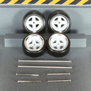May include: Four black model car tires with white rims and silver accents. Also included are several silver metal rods and pins, likely for assembly. The tires have a four-spoke design.