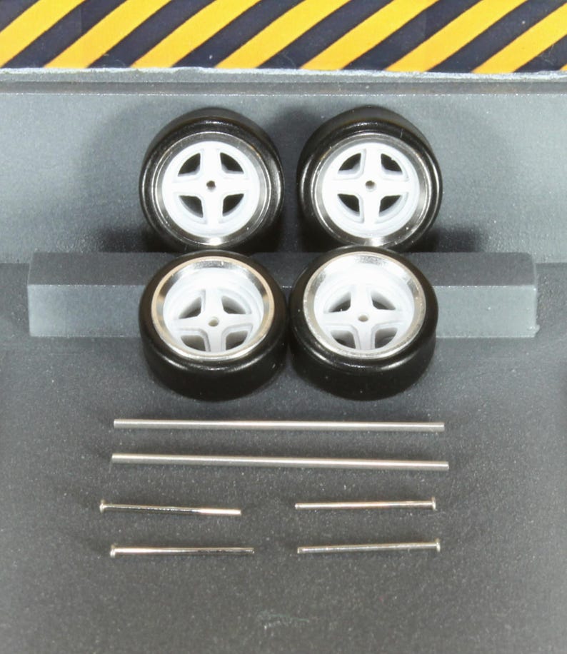 May include: Four black model car tires with white rims and silver accents. Also included are several silver metal rods and pins, likely for assembly. The tires have a four-spoke design.