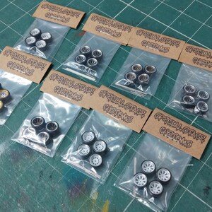 May include: Several sets of model car wheels, each in a clear plastic bag with a brown label. The wheels feature different designs, including black and silver rims, and white rims. The labels display "Special Stage Customs".