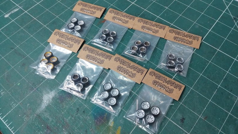 May include: Several sets of model car wheels, each in a clear plastic bag with a brown label. The wheels feature different designs, including black and silver rims, and white rims. The labels display "Special Stage Customs".