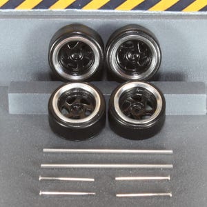 May include: Four black model car wheels with silver rims and a five-spoke design. Several silver metal rods are also present. The wheels are arranged in a square formation, suggesting a set for a miniature vehicle.