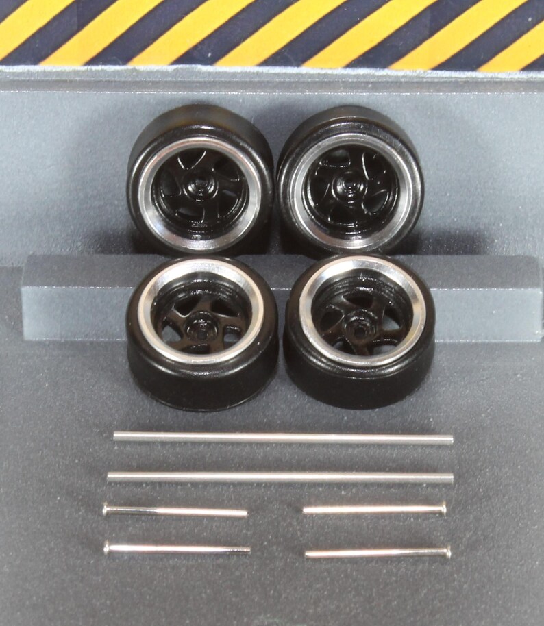 May include: Four black model car wheels with silver rims and a five-spoke design. Several silver metal rods are also present. The wheels are arranged in a square formation, suggesting a set for a miniature vehicle.