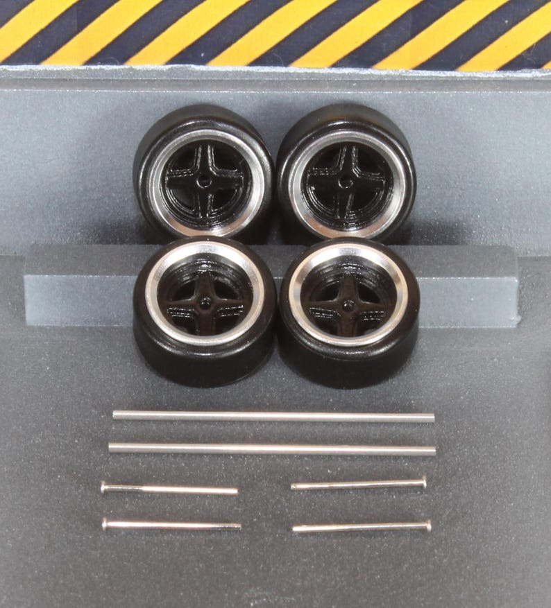 May include: Four black model car wheels with silver rims and a cross-shaped spoke design. Also included are four silver metal rods and two silver metal pins, likely for assembly. The wheels are arranged in a square.