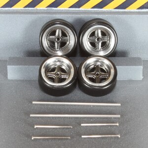 May include: Four black model car tires with silver rims and a cross-shaped spoke design. Also included are several silver metal rods of varying lengths, likely for model car assembly. The tires are set against a gray background.