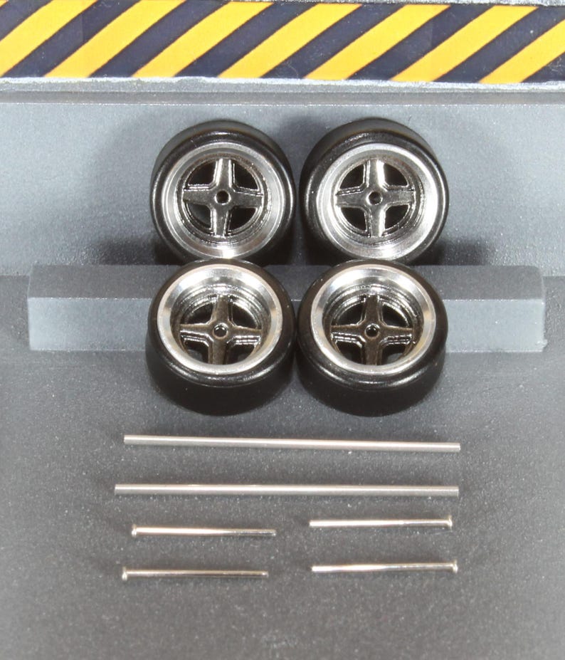 May include: Four black model car tires with silver rims and a cross-shaped spoke design. Also included are several silver metal rods of varying lengths, likely for model car assembly. The tires are set against a gray background.