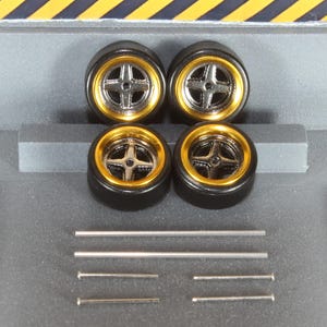 May include: Four black model car wheels with gold rims and silver cross-shaped spokes. Several silver metal rods and pins are also included, likely for assembly. The wheels are set against a gray background with yellow and black diagonal stripes.