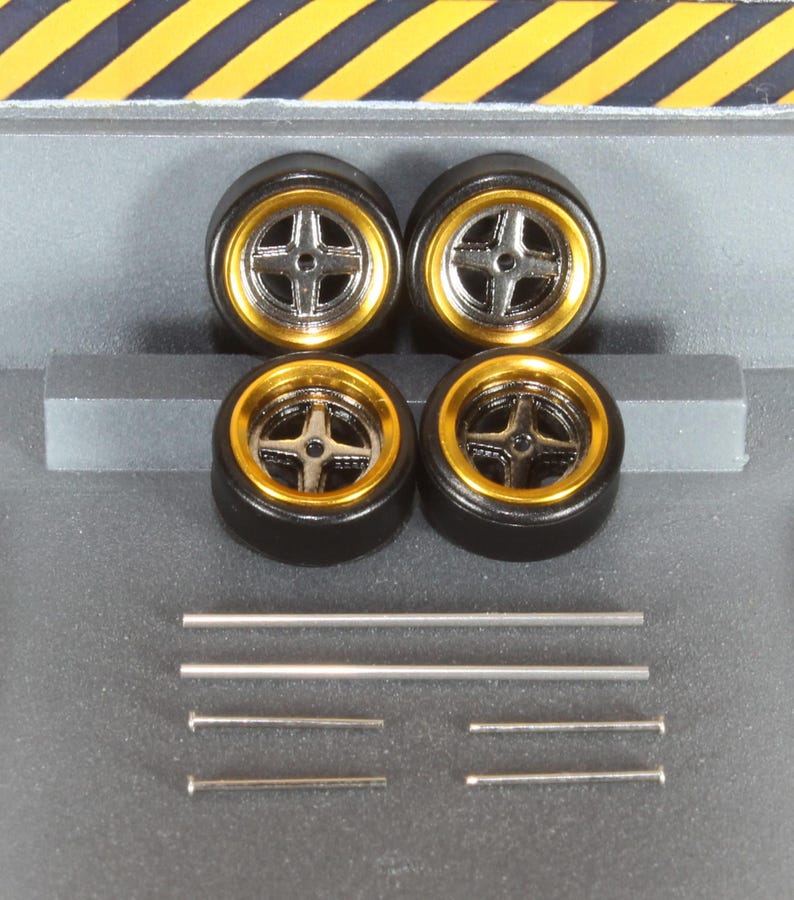 May include: Four black model car wheels with gold rims and silver cross-shaped spokes. Several silver metal rods and pins are also included, likely for assembly. The wheels are set against a gray background with yellow and black diagonal stripes.
