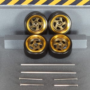 May include: Four black model car wheels with gold rims and silver spokes. The wheels are arranged in a square, with several silver rods and pins below them. The background is grey with yellow and black diagonal stripes.