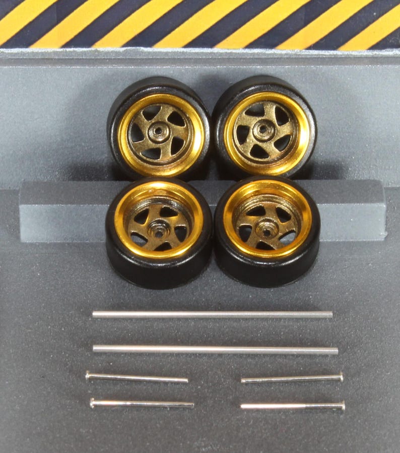 May include: Four black model car wheels with gold rims and silver spokes. The wheels are arranged in a square, with several silver rods and pins below them. The background is grey with yellow and black diagonal stripes.