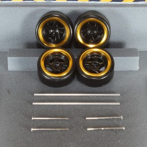 May include: Four black model car wheels with gold rims are shown. The wheels are arranged in a square, with several silver metal rods and pins below them. The background features a grey surface with yellow and black diagonal stripes.