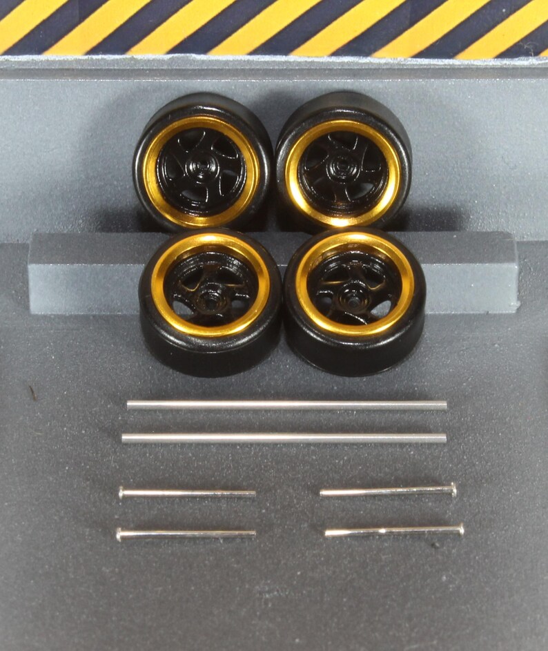 May include: Four black model car wheels with gold rims are shown. The wheels are arranged in a square, with several silver metal rods and pins below them. The background features a grey surface with yellow and black diagonal stripes.