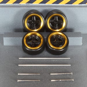 May include: Four black model car wheels with gold rims and silver axles. The wheels have a five-spoke design. The axles are of varying lengths. The background is a gray surface with yellow and black diagonal stripes.