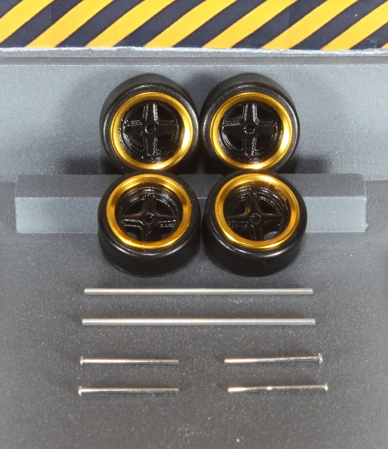 May include: Four black model car wheels with gold rims and silver axles. The wheels have a five-spoke design. The axles are of varying lengths. The background is a gray surface with yellow and black diagonal stripes.