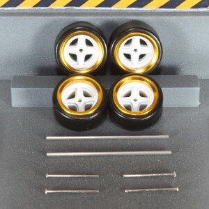 May include: Four black model car tires with white and gold rims. The tires are arranged in a square, with several silver metal rods and pins below them. The background is gray with yellow and black diagonal stripes at the top.