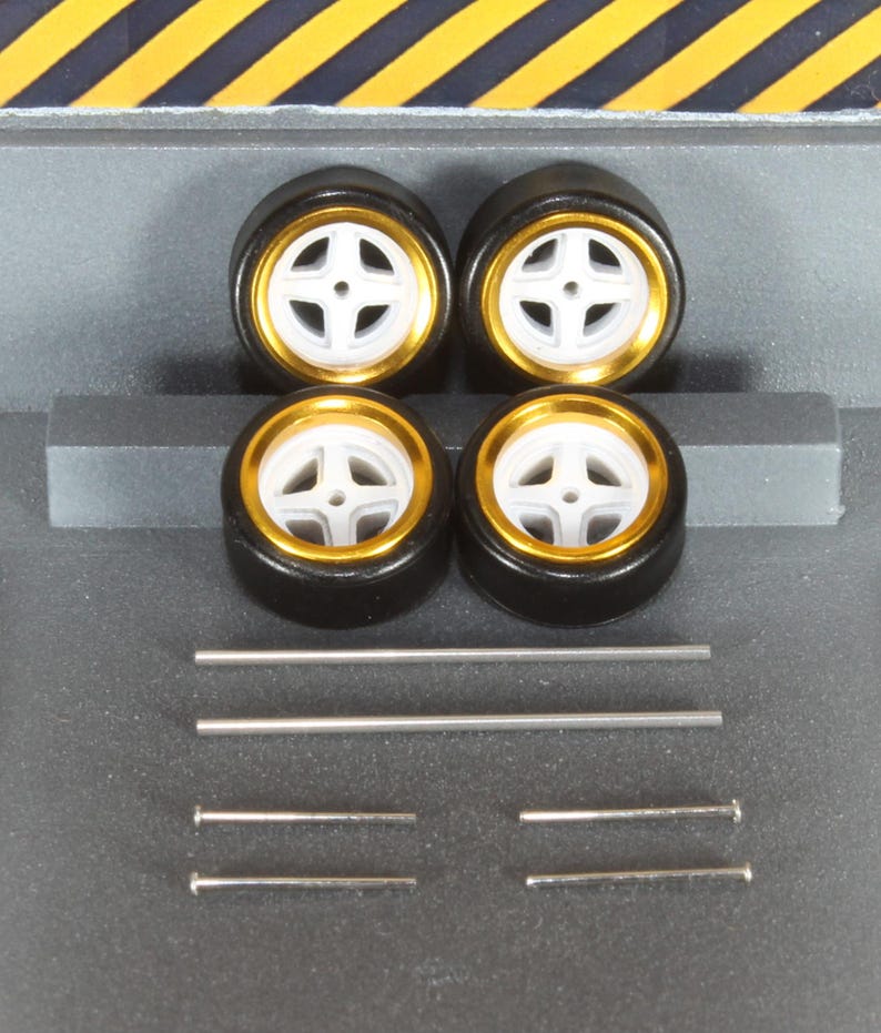 May include: Four black model car tires with white and gold rims. The tires are arranged in a square, with several silver metal rods and pins below them. The background is gray with yellow and black diagonal stripes at the top.
