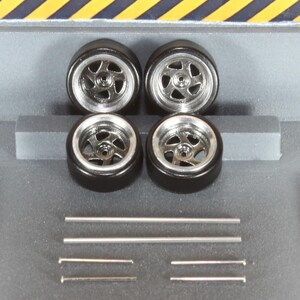 May include: Four black model car tyres with silver rims and several silver metal rods. The tyres have a five-spoke design. The background is a grey surface with yellow and black diagonal stripes.