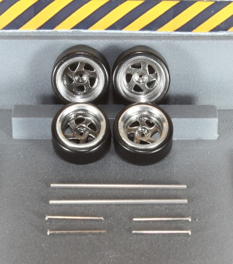 May include: Four black model car tyres with silver rims and several silver metal rods. The tyres have a five-spoke design. The background is a grey surface with yellow and black diagonal stripes.