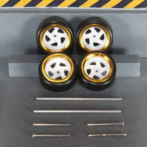 May include: Four model car wheels with black tyres, gold rims, and white spokes. Several silver metal axles and pins are also included. The wheels are arranged in a square, with the axles and pins below.