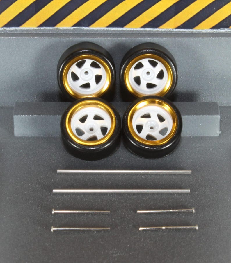 May include: Four model car wheels with black tyres, gold rims, and white spokes. Several silver metal axles and pins are also included. The wheels are arranged in a square, with the axles and pins below.