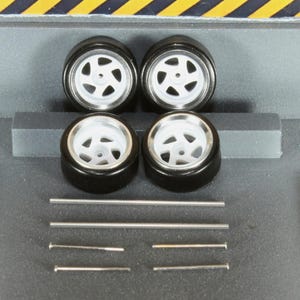 May include: Four black model car tyres with white rims and silver spokes. Several silver metal rods and pins are also included, likely for assembly. The tyres are arranged in a square, suggesting a set for a miniature vehicle.