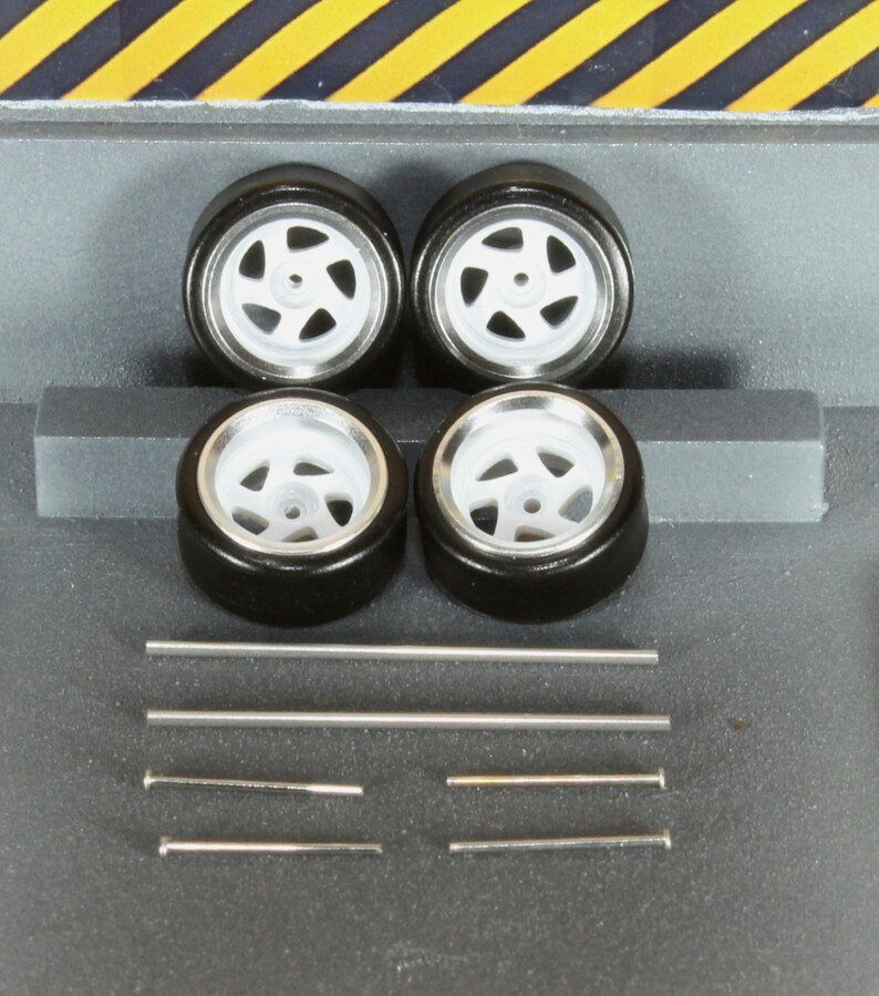 May include: Four black model car tyres with white rims and silver spokes. Several silver metal rods and pins are also included, likely for assembly. The tyres are arranged in a square, suggesting a set for a miniature vehicle.