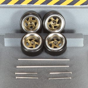 May include: Four model car wheels with black tyres, gold-coloured centres, and silver rims. Several silver metal rods of varying lengths are also present, possibly for axles or detailing.
