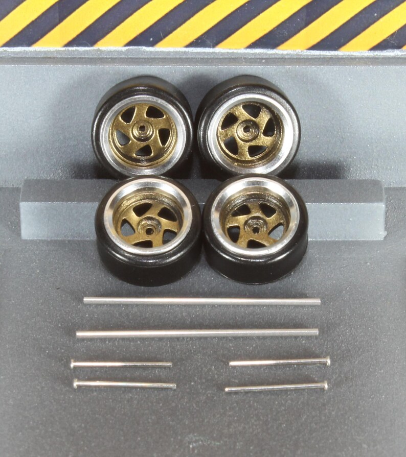 May include: Four model car wheels with black tyres, gold-coloured centres, and silver rims. Several silver metal rods of varying lengths are also present, possibly for axles or detailing.