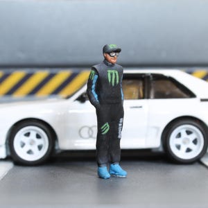 Puede incluir: Una figura en miniatura de un piloto de carreras con un traje de carreras negro con detalles verdes y azules, una gorra y gafas de sol. La figura está de pie frente a un coche deportivo blanco. El traje de carreras tiene el logo de Monster Energy.