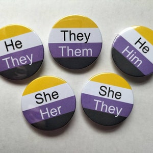 May include: Five round buttons featuring the colours of the non-binary pride flag. The buttons display different text, including "He They", "They Them", "He Him", "She Her", and "She They".