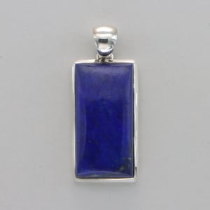 May include: A rectangular pendant featuring a deep blue stone set in a silver-colored frame. The stone has a polished surface and is a rich, solid blue color. The pendant has a loop at the top for a chain.