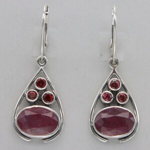 May include: A pair of silver dangle earrings with a teardrop shape. Each earring features a large oval-shaped pink gemstone surrounded by four smaller round red gemstones.