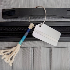 May include: A gray suitcase with a black handle and a white luggage tag attached to it with a blue and white braided cord. The tag has the word "Name" printed on it.