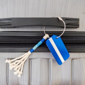 May include: A blue luggage tag with a white label that says "Name" attached to a gray suitcase with a black handle. The tag is connected to the suitcase with a silver chain and a white and blue braided rope.