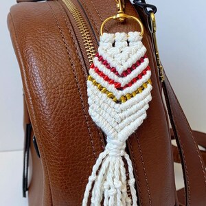 May include: A white macrame keychain with a red, white, and gold striped pattern and a white tassel attached to a gold key ring. The keychain is attached to a brown leather backpack.