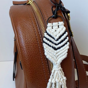 May include: A white and black macrame keychain with a tassel attached to a brown leather backpack.