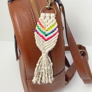 May include: A white macrame keychain with a rainbow colored design and a tassel. The keychain is attached to a brown leather backpack.